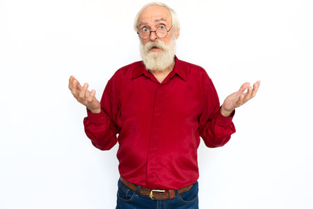 Confused senior man shrugging. Surprised Caucasian male model with gray hair and beard in red shirt and glasses looking at camera with raised hands, showing helpless gesture. Confusion, choice conceptの写真素材