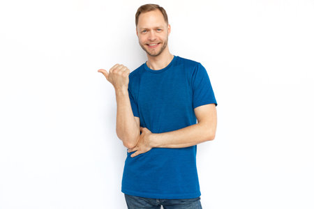 Cheerful man pointing thumb at copyspace. Fair hair Caucasian man in T-shirt looking at camera while standing on white background and promoting product. Advertisement, promotion, conceptの写真素材