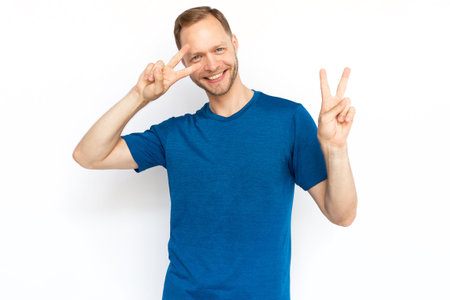 Happy Caucasian man showing peace sign at camera. Bearded man in T-shirt looking at camera, smiling and standing on white background. Satisfaction, achievement, winning conceptの写真素材