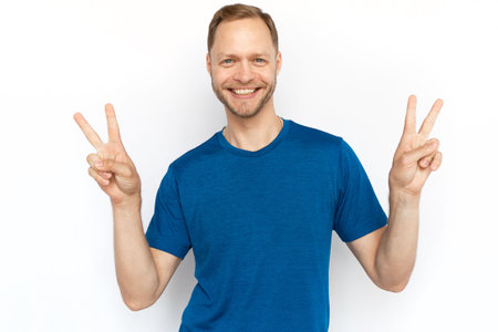 Caucasian man showing peace signs at camera. Bearded man with fair hair looking at camera, standing on white background and smiling. Satisfaction, achievement, winning conceptの写真素材