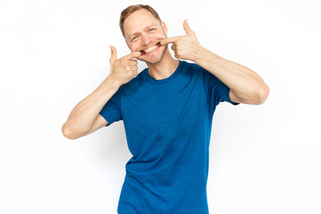 Caucasian man making fake smile by his fingers. Portrait of bearded man looking at camera on white background, showing fake emotions. Expression, feeling, negative emotions conceptの写真素材