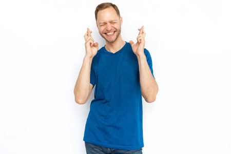 Excited man with crossed fingers standing on white background. Happy Caucasian guy praying for success, squeezing eyes, smiling, hoping for luck. Hope, wish, superstition conceptの写真素材