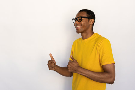 Portrait of happy young man wearing eyeglasses showing approval sign over white background. African American guy wearing yellow T-shirt looking away and making thumbs up. Satisfaction conceptの写真素材