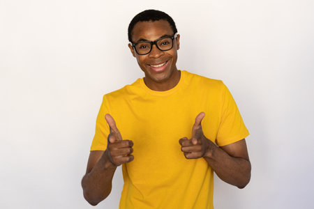 Portrait of happy young man wearing glasses making thumbs up over white background. African American guy wearing yellow T-shirt showing like gesture and smiling at camera. Approval conceptの写真素材