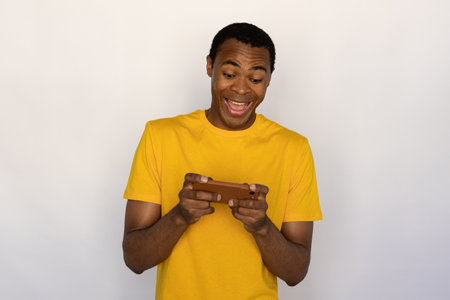 Portrait of excited young man watching video on mobile phone over white background. African American guy wearing yellow T-shirt surfing internet on smartphone. Mobile technology conceptの写真素材
