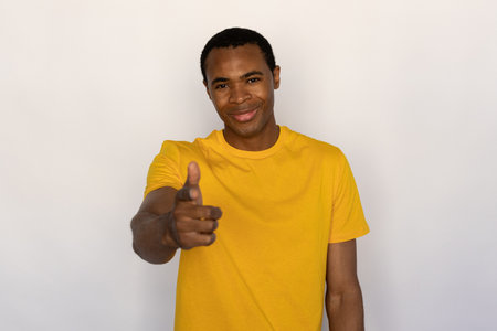Portrait of young man pointing at camera and sneering against white background. African American guy wearing yellow T-shirt looking at camera and smiling. Mockery conceptの写真素材