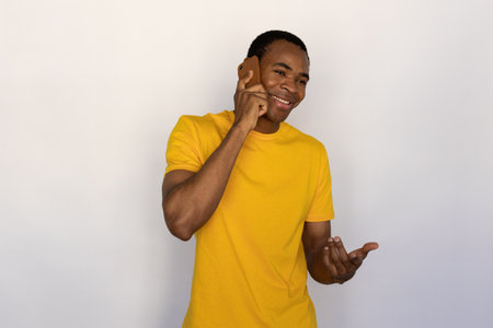 Portrait of happy young man having mobile conversation with friend over white background. African American guy wearing yellow T-shirt talking on smartphone and smiling. Mobile communication conceptの写真素材