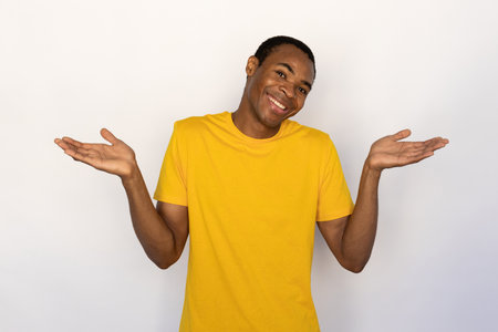 Portrait of happy young man shrugging shoulders over white background. African American guy wearing yellow T-shirt expressing indifference. Bewilderment conceptの写真素材