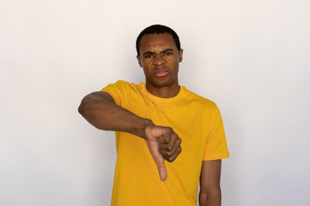 Portrait of displeased young man making dislike gesture against white background. African American guy wearing yellow T-shirt showing thumb down. Disapproval conceptの写真素材