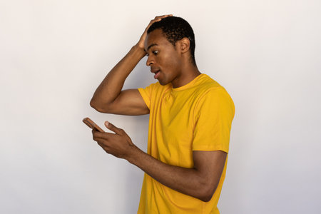 Portrait of shocked young man reading message or newsletter on white background. African American guy wearing yellow T-shirt looking at mobile phone in stress. Mobile communication and stress conceptの写真素材