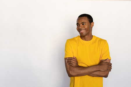 Portrait of happy young man posing with folded arms against white background. African American guy wearing yellow T-shirt looking away and smiling. Happiness conceptの写真素材
