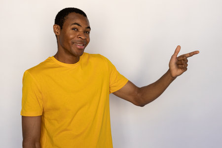 Portrait of happy young man pointing aside and smiling over white background. African American guy wearing yellow T-shirt advertising something. Promotion conceptの写真素材