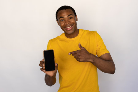 Portrait of happy young man pointing at mobile phone over white background. African American guy wearing yellow T-shirt advertising new model of smartphone and smiling at camera. Promotion conceptの写真素材