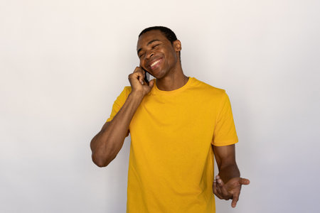 Portrait of positive young man talking on smartphone with closed eyes over white background. African American guy wearing yellow T-shirt calling friend with mobile phone. Mobile communication conceptの写真素材
