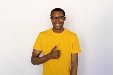 Portrait of cheerful young man wearing glasses showing like gesture over white background. African American guy wearing yellow T-shirt making thumb up and smiling at camera. Approval conceptの写真素材