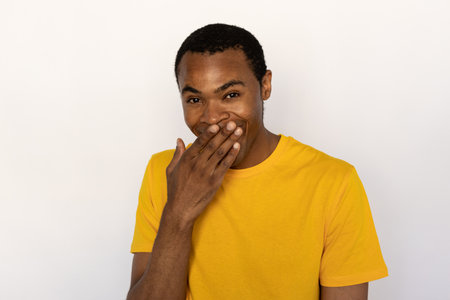 Portrait of positive young man sneering and covering mouth with hand over white background. African American guy wearing yellow T-shirt looking at camera and smiling. Mockery conceptの写真素材