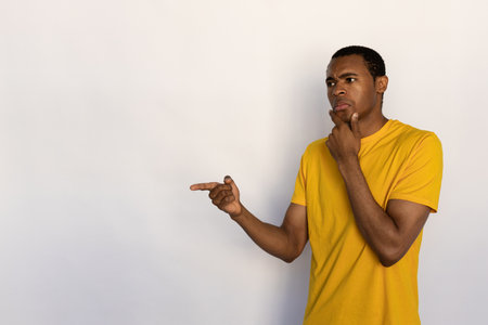 Portrait of pensive young man pointing aside against white background. African American guy wearing yellow T-shirt showing something with thoughtful expression. Doubt and uncertainty conceptの写真素材