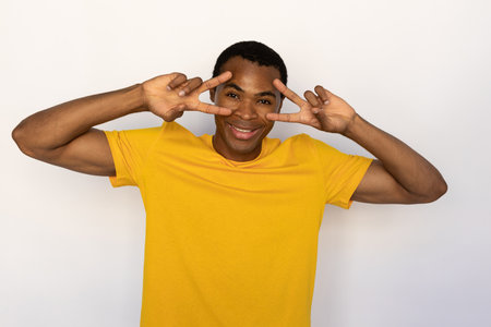 Portrait of happy young man making victory gesture over white background. African American guy wearing yellow T-shirt showing v-sign at eyes and smiling at camera. Fun and happiness conceptの写真素材