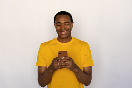 Portrait of cheerful young man using mobile phone. African American guy wearing yellow T-shirt reading message on mobile phone and smiling over white background. Mobile communication conceptの写真素材