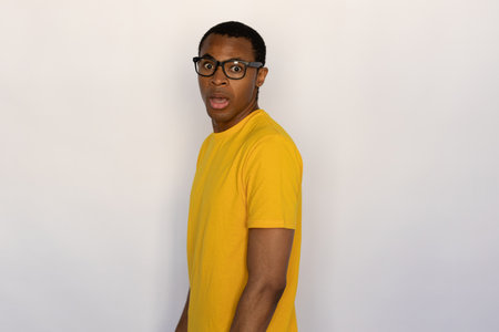 Terrified young man opening his mouth from shock. Male African American model with short black haircut in black eyeglasses wearing yellow T-shirt seeing something scary. Emotion, fright conceptの写真素材