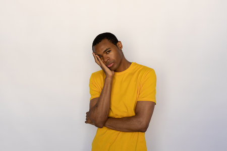Bored young man holding his face with hand. Male African American model with brown eyes and short black haircut in yellow T-shirt looking bored listening to something uninteresting. Boredom conceptの写真素材