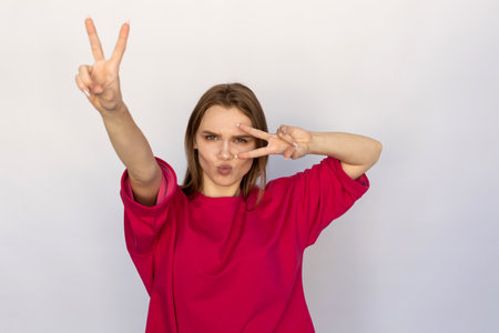 Winsome young woman showing V-signs. Caucasian female model with brown hair and green eyes in pink...