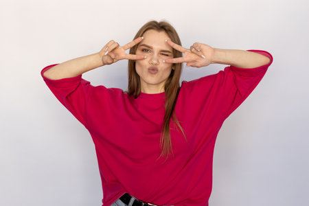 Cheerful young woman showing V-signs near eyes. Caucasian female model with brown hair and green eyes in pink sweater showing victory signs, winking and puckering lips. Coquetry, flirt conceptの写真素材