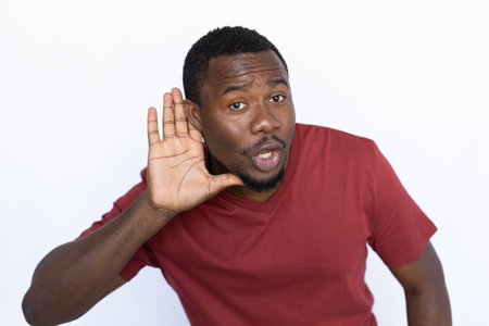 Listening young man with hand over ear. Male African American model with brown eyes, short black haircut and beard in maroon T-shirt listening to rumor or gossip. Deafness, silence concept.の写真素材