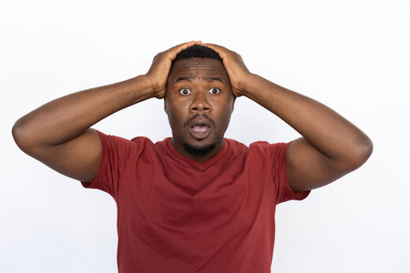 Amazed young man holding his hands on head from shock. Male African American model with brown eyes, short black haircut and beard in maroon T-shirt opening his mouth from surprise. Amazement conceptの写真素材