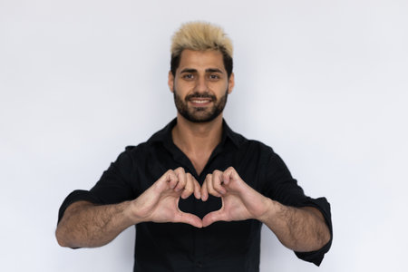 Close-up of heart gesture made by joyful businessman. Male Caucasian model with brown eyes, ombre painted hair and beard in black shirt showing heart sign with his hands. Love, feeling conceptの写真素材