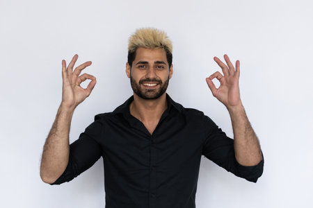 Cheerful businessman showing OK gesture with both hands. Male Caucasian model with brown eyes, ombre painted hair and beard in black shirt smiling assuring everything OK. Confidence conceptの写真素材