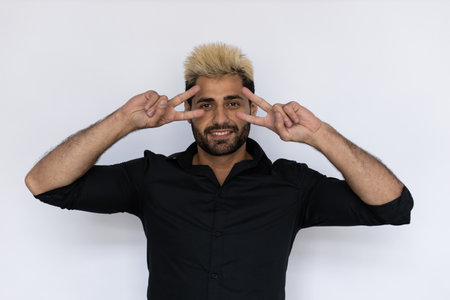 Winsome young man showing V-signs near his eyes. Male Caucasian model with brown eyes, ombre painted hair and beard in black shirt smiling making victory signs. Self-assurance conceptの写真素材