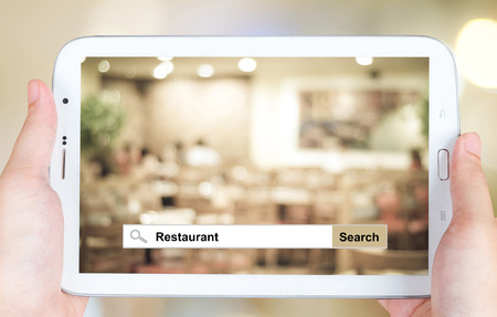 Hand holding tablet with restaurant word on search bar over blurred restaurant background, restaurant reservation, food online, food delivery conceptの写真素材