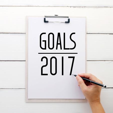 Hand holding pen over  paper clipboard with 2017 goals on white wood background, sucess in business  conceptの写真素材