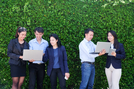 Business group of people with computer labtop, tablet, smart phone planning their work standing in front of green tree leaves backgroundの写真素材