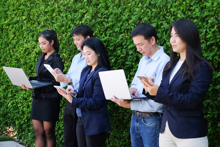 Business group of people with computer labtop, tablet, smart phone planning their work standing in front of green tree leaves backgroundの写真素材