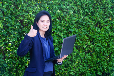 Business woman holding lab-top computer and thumbs up gesture standing in front of green tree leaves background, business success conceptの写真素材