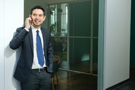 Business man talking smart phone with happy smiling face standing in front of meeting room inside office building background, businessman on smart phone, on line conference conceptの写真素材