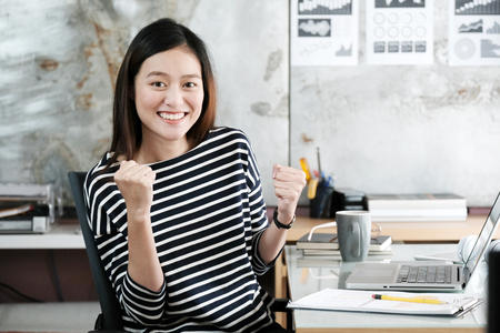 Young businesswoman with arms up and smiling face while working with laptop computer at her office table, success in business conceptの写真素材
