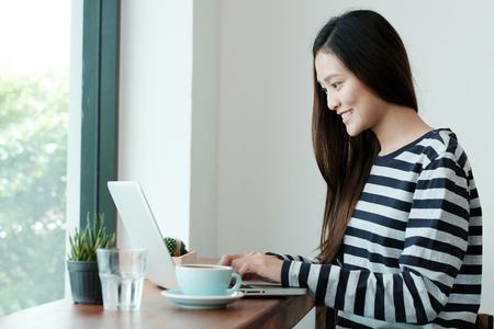 Beautiful asian woman using laptop computer while sitting by window, work at home, people education, business and technology, casual lifestyle conceptの写真素材