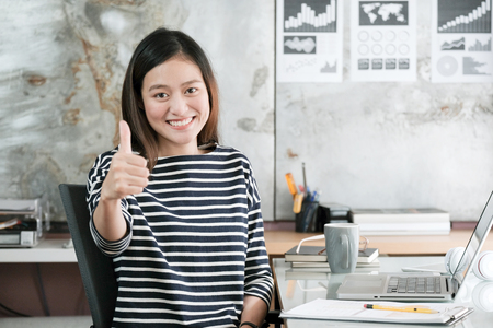 Young asian businesswoman thump up and smiling while sitting at her desk office, positive expression, sucess in business conceptの写真素材