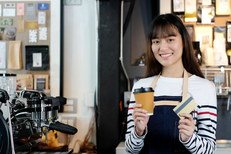 Young asian woman barista holding a disposable coffee cup and credit card with smiling face at cafe counter background, small business owner, food and drink industry conceptの写真素材