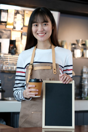 Young asian woman barista holding blank chalkboard with smiling face at cafe counter background, food and drink conceptの写真素材