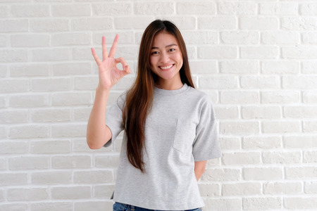 Portrait of young asian woman showing ok hand sign and smiling while standing over white brick wall background, people with positive gestureの写真素材