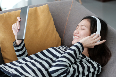 Young asian woman wearing headphones and holding tablet smiling with happiness on sofa, lifestyle conceptの写真素材
