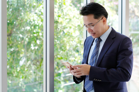Asian Businessman holding smart phone with happy smiling face standing by windows, inside eco office building background, businessman on smart phoneの写真素材