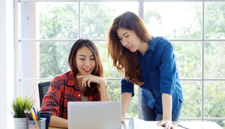 Two young asian women working with laptop computer at home office with happy emotion moment, working at home, small business, office casual lifestyle conceptの写真素材