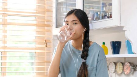 Young beautiful asian woman drinking water while standing by window in kitchen background, people...