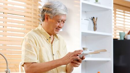 Senior asian man using smartphone at home background, Happy retirement asia male holding phone while standing in kitchen, Active senior people and technology lifestylesの写真素材