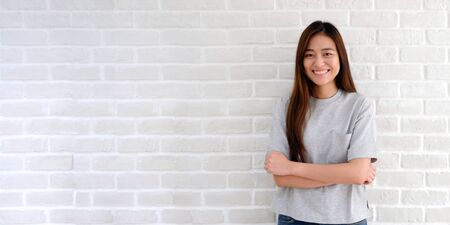 Portrait of young asian woman standing and smiling over white brick wall background with copy space, Happy asia girl smiling and looking at camera in casual lifestyleの写真素材